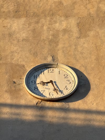 A close-up photograph taken during daylight hours captures a beige-toned wall with a white-faced clock mounted on it. The clock, displaying the time as approximately 8:25, casts a distinct shadow on the wall, indicating a direct light source. The wall's surface has a rough, textured appearance. A vertical shadow line also cuts across the right side of the image. The name "F. VAQUER" is visible on the clock face near the 7 and 8 o'clock positions. The image location could be determined based on that information, and Vinaròs is the location.