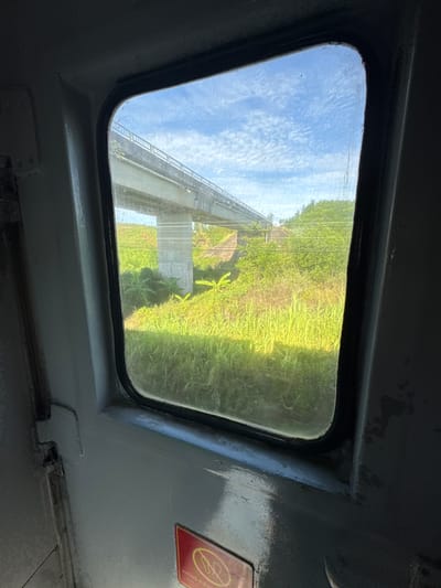 An exterior view from a train window shows a bright, cloudy sky and a lush green landscape. A concrete overpass spans across the middle ground, supported by concrete pillars. The landscape below consists of tall, dense green grass and various leafy plants. In the foreground, the interior of the train is visible, with gray painted walls, a black window frame, and a red warning sticker with Vietnamese text.