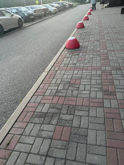 A row of red bollards with white tops are lined up on a patterned brick walkway. The walkway is made of rectangular bricks in alternating shades of grey and pink. To the left of the walkway is a paved area with a row of parked cars. The cars are of various colors including white, grey, and black. The scene appears to be outdoors, likely a street or parking lot. The lighting suggests it is daytime. There is a slight tilt to the image, making it appear as if it was taken from a slightly elevated or angled perspective. No text is clearly visible in the image.