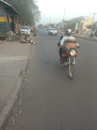 The image shows a street scene in New Karu, Nigeria (latitude: 8.998, longitude: 7.571). There is a man driving a motorcycle with another person riding as a passenger. The motorcycle has a large, brown container strapped to the back. The motorcycle is driving on a paved road in the middle of the image, heading towards the right side of the image. There are buildings on the right side of the road and trees on the left. The scene is taken during the day. There is a parked car on the left side of the road, near a sidewalk. There is a building on the left side of the image. There are traffic cones and a tire on the sidewalk. There are two people walking on the sidewalk on the left side of the image. The sky is a light gray. The ground is covered in asphalt and there are patches of dirt. The image appears to be taken from a distance and angled slightly downwards.