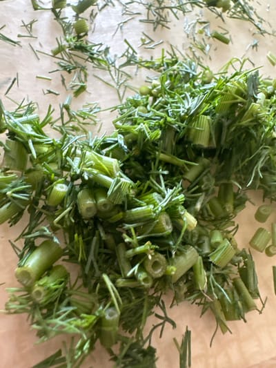 The image shows a pile of chopped dill weed and stems on a light-colored surface, possibly parchment paper. The dill is vibrant green and appears fresh. The stems are cut into short, cylindrical pieces, some showing hollow centers. The dill fronds are finely cut and mixed throughout the stems. The lighting suggests it might be daytime, but there are no specific cues for time of day or weather. The setting appears to be a kitchen or food preparation area. There is no visible text in the image. The focus is entirely on the prepared dill.