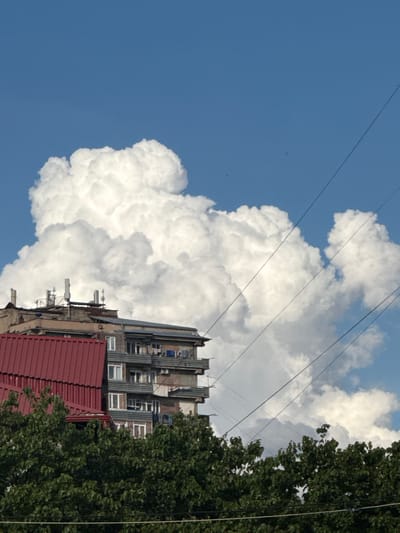 The image depicts a cloudy sky above a multi-story building in Abovyan, Armenia. The building has a red-tiled roof in the foreground, with an adjacent concrete structure featuring multiple balconies with laundry hanging. Several antennas and communication towers are visible on the rooftop of the building. Dark green trees line the bottom of the frame, partially obscuring the lower levels of the buildings. Several power lines stretch diagonally across the sky. The sky is a bright blue, with large, white cumulus clouds dominating the upper portion of the image. There are no people visible in the scene.