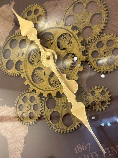 The image shows a close-up view of a decorative clock with a steampunk aesthetic. The clock features multiple brass-colored gears of various sizes arranged in an overlapping pattern. A single brass clock hand, pointed and decorative, extends from the center. The background appears to be a dark brown surface with a faint, textured map design and some text that reads "1867" and "ARD TIM". There are also several small, bright circular reflections on the surface, possibly from lighting. There are no people or animals in the image, nor any indication of a specific location in Deventer, Netherlands.