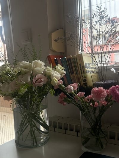 The image shows a still life of flowers and books in an indoor setting. Two bouquets in clear glass vases are positioned in front of a window. The bouquet on the left features white and pink flowers with delicate green foliage, while the bouquet on the right primarily consists of pink flowers. Between the bouquets, a stack of books is visible. The book titles and authors include "45 татуировок личности" by Максим Батырев, "Психолог" by Т.И. - сама себе, and "Эмили Бронте" by Sarah Ada. The background shows a blurred view of buildings through the window. Given that the books are in Russian, and the books feature Serbian author and a Serbian book (КАРЛ ГУСТАВ ЈУНГ), and the general style, the location is likely in a country where Russian literature is read, such as Serbia, and given that the books were purchased, the location is likely a major city like Novi Sad, Serbia.