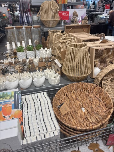 This image displays a retail store setting, likely a home decor or gift shop in Erlenbach am Main, Germany. The scene is filled with various decorative items, primarily made of natural materials like woven straw and jute. Prominently featured are large woven baskets, one circular and another cylindrical, along with smaller woven cylindrical holders that appear to be candle holders or decorative covers.
Interspersed among the woven items are white ceramic decorations, including small house-shaped ornaments and crown-shaped holders with intricate cut-out patterns. There are also glass jars with wooden lids, containing dried decorative elements and small potted plants with green foliage. Other items visible include stacks of white napkins with black stripes, and a colorful picture of oranges in a glass.
The presence of price tags and discount signs ("15% auf Dekoration") indicates a commercial environment where these items are for sale. The lighting suggests it is daytime, likely during business hours, as there is a hint of ambient store lighting. While no people are clearly visible, the blurred background suggests other shoppers or displays typical of a retail store. The overall impression is one of a well-stocked store offering rustic and decorative home accessories.