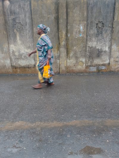 In Agani, Nigeria, a single adult female is observed walking from left to right on a damp, dark paved road. She is dressed in a patterned teal, blue, and yellow traditional garment, including a matching headwrap. Her left arm is slightly bent, and she carries an orange fabric bag in her right hand. Her footwear consists of brown shoes or sandals. The background features a weathered concrete or stucco wall with vertical indentations and two star-shaped decorative medallions. The road surface exhibits some light brown streaks or deposits. The lighting is diffused.