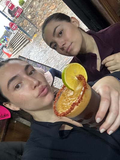 Two adult females are captured in a selfie-style photograph, appearing to be seated indoors or in a covered patio area in Huasca de Ocampo, Mexico. The foreground subject, on the left, has dark hair pulled back, light brown eyes, and is making a pouting expression towards the camera. She wears a black garment with cutouts near the collarbone and holds a tall, dark glass with her right hand. The glass contains a dark brown beverage with a frothy, lighter brown head, and its rim is heavily coated with a reddish-brown spice mixture. A thick slice of lime or orange is garnished on the rim. The background subject, on the right, also has dark hair pulled back and wears a dark purple collared shirt with a subtle silver necklace; she looks directly at the camera with a neutral expression.

Through an opening behind the subjects, an outdoor scene is visible. A prominent stone wall, constructed from irregular gray and reddish-brown stones, forms the primary background structure. Multiple signs are affixed to a pole in the background. A red sign with white text indicates "El Zócalo" and "Comedor Tradicional," followed by "Ruta de los [Prismas Basálticos]" and "Huasca de Ocampo," unequivocally identifying the city. Another red sign displays "1x1," potentially indicating a promotional offer. Stone steps lead downwards to the left of the stone wall, and part of a light-colored building with a column is visible in the upper left background. The overall lighting suggests overcast daylight outdoors.