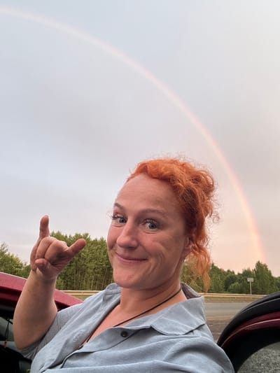 A woman with bright red, curly hair is captured in a selfie taken from a low angle. She is wearing a light blue button-up shirt. Her right arm is bent, and she is making a "shaka" hand gesture, with her thumb and pinky extended. She is smiling at the camera.
Behind her, a faint rainbow arcs across a pale, overcast sky. The background shows a line of trees and a hint of a road with a street sign visible. The edge of a car, a dark red color, frames the right side of the image. The lighting suggests it might be late afternoon or early evening, with the overcast sky creating a soft, diffused light. The presence of the rainbow indicates recent or ongoing rain. The location is identified as Sovkhoznyi, Russia.