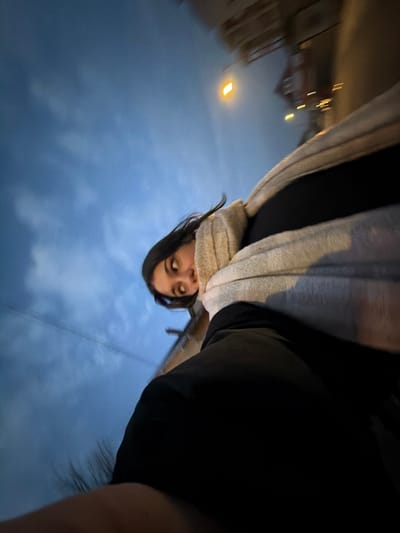 An off-kilter, upward-angled selfie captures a person with dark hair and a beige scarf, set against a dusk sky. Streetlights glow in the blurred background, suggesting movement. The person's gaze is directed at the camera, with an expression that could be interpreted as quizzical or slightly surprised. A dark garment is visible in the foreground, obscuring some of the sky. The overall composition feels casual and immediate, like a spontaneous moment captured.