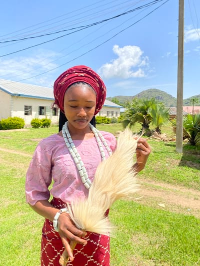 A young woman is photographed outdoors, wearing traditional attire. She has a red headwrap adorned with a gold pattern and a pink lace top. Around her neck, she wears multiple strands of white beads, and a bracelet of the same beads is on her wrist. In her hands, she holds a whisk-like object made of blonde, feathery material, with a wooden handle. Her eyes are closed, and she has a serene expression.

The setting appears to be in Vom, Nigeria. The background shows a building with a white facade and windows, indicative of an institutional or residential structure. Lush green grass covers the ground, with some bushes visible. In the distance, a hill or mountain can be seen under a bright blue sky with scattered white clouds. A utility pole and power lines are present in the foreground, suggesting the presence of infrastructure. The lighting indicates it is daytime, likely late morning or early afternoon, with bright sunlight casting shadows. The overall atmosphere is peaceful and evokes a sense of cultural tradition. No visible text is present in the image.