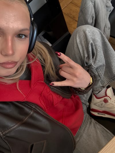 The image captures a rotated, close-up perspective of a person indoors, likely in Antwerp, Belgium. The main subject, positioned with their head towards the bottom left, is a young individual with blonde hair, seen wearing black over-ear headphones. Their face is partially visible, showing a brown eye with defined lashes, the bridge of their nose, and a slight hint of a smile.
They are dressed in a vibrant red hoodie layered beneath a dark brown leather jacket. A gold-toned watch adorns their wrist, and their hand is making a "rock on" or "devil horns" gesture, with nails painted in a distinctive black and red pattern.
In the upper portion of the frame, the lower body of another person, or potentially the same individual, is visible. This person is wearing light grey, acid-wash style jeans and white sneakers with prominent red accents, appearing to be Air Jordan 4s. The background provides glimpses of a dark, possibly wooden or upholstered, surface and a light brown wooden floor, indicative of a casual indoor setting such as a cafe or lounge. The overall atmosphere conveyed is relaxed and informal. No text is visible within the image.
