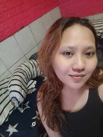 A close-up, selfie-style photograph is presented, focusing on the face of a young woman with medium-toned skin and reddish-brown hair. She is wearing a black tank top. Her expression is neutral to slightly smiling. The background consists of a bed with striped pillows and a dark blue sheet with white stars. A red textured wall is visible in the upper left portion of the image. The overall lighting is warm and appears to be artificial. The image was taken indoors, likely in a bedroom. Based on the context, the location is the city of Depok, Indonesia.