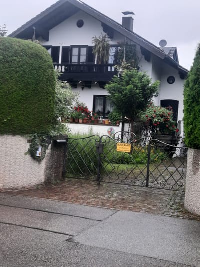 The image captures a view of a white, two-story house with a dark, pitched roof and a black chimney in Rosenheim, Germany. A black satellite dish is visible on the roof. The house features dark shutters on its windows and a black balcony with decorative railings. Hanging plants adorn the balcony and the front of the house. In front of the house, a green, manicured hedge is prominent on the left, and another tall green evergreen tree is on the right. A decorative black wrought-iron gate with an arched top and diamond-shaped accents is in the foreground, leading to a paved driveway. A yellow sign with German text reading "AUSFAHRT freihalten Bitte auch gegenüber" is attached to the gate. The driveway is paved with red brick pavers that appear wet, suggesting recent rain. A portion of the street in front of the driveway is also visible, paved with asphalt and exhibiting water. A stone wall with a textured finish borders the left side of the driveway and the hedge. Several potted plants with red flowers are arranged on a ledge below the balcony.