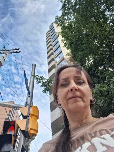 This is a selfie taken outdoors in a city, likely Ottawa, Canada. The subject is a middle-aged woman with brown hair, looking directly at the camera. She is wearing a beige sweatshirt with large white lettering that is partially visible. She has hoop earrings on both ears.
In the background, there is a tall, modern building with alternating light and dark sections, characteristic of some architectural styles found in Canadian cities. To the left, there is a traffic light, currently displaying a red "walking person" symbol, indicating that it is not safe to cross. A utility pole with numerous wires is also visible, along with a blue street sign. A large tree with dense green foliage is present on the right side of the frame, partially obscuring the building. The sky is cloudy with patches of blue. The perspective of the selfie is from a low angle, looking upwards.
