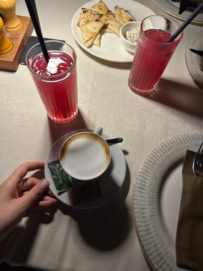 A overhead view of a table setting shows two glasses of red beverage with straws, a cup of coffee with foam, a plate of flatbread with a small bowl of white dip, two packets of sugar, and a fork and napkin on a white tablecloth. The scene appears to be in Bartula, Montenegro, with the ambient lighting suggesting an evening setting.