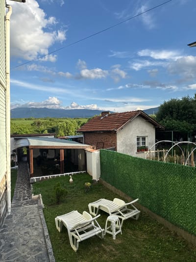 The image depicts a residential backyard in Ivanyane, Bulgaria. The scene is bathed in natural light, with a partly cloudy blue sky overhead. A two-story house with a red tiled roof is visible, adjacent to a smaller, single-story structure with a similar roof and white stucco walls. A covered patio area with glass enclosure is also present. In the foreground, two white plastic lounge chairs and a small white plastic stool are arranged on a grassy lawn. A decorative goose statue stands near the patio. A stone pathway leads from the left edge of the frame towards the patio. A dense green hedge forms the boundary of the backyard to the right, behind which a white arched structure, possibly a garden hoop house, and some plants are visible. In the distance, lush green fields and hills can be seen, with mountains forming the backdrop. The exterior of a light green, textured wall of a building occupies the far left of the frame.