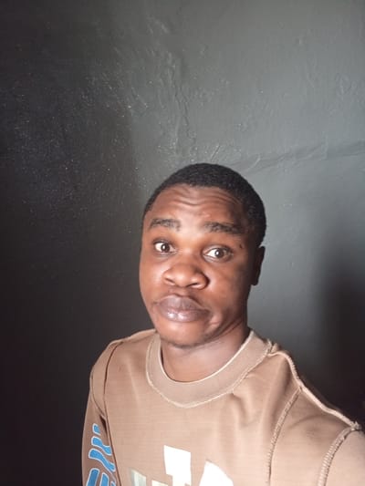 A frontal portrait depicts an adult male, of African descent, with dark skin, short dark hair, and brown eyes. He wears a light brown, long-sleeved crewneck shirt featuring prominent external stitching along the shoulders and neckline. A partial design in light blue and white is visible on the viewer's right sleeve. The subject gazes directly into the camera with a neutral expression and slightly parted lips. The background consists of a uniformly dark, textured wall, exhibiting subtle surface variations and minor lighter speckling. This scene is located in the city of Owerri, Nigeria.