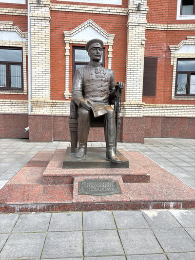 This is a vertical, eye-level, full shot of a bronze statue of a man sitting on a bench, holding a rifle. The statue is located outdoors, in front of a modern brick building. The man is wearing a military uniform with a hat and medals. He is depicted in a relaxed posture, with his legs crossed and his rifle resting on his lap.

The building behind the statue is made of red brick with white decorative elements. There are windows visible on the building. The ground around the statue is paved with granite tiles. A plaque with text is attached to the granite base of the statue.

The plaque reads: "АЛЕКСАНДР АНДРЕЕВИЧ КЕЛЛЕР". This indicates the statue is of Aleksandr Andreevich Keller.

The scene appears to be set in a public space, possibly a city square or a memorial site. The lighting suggests it is daytime, and the weather appears to be overcast, as there are no strong shadows. The overall mood is somber and respectful, befitting a monument.

The statue is a prominent feature of the scene, drawing the viewer's attention. The details of the uniform and the rifle are rendered with precision. The surrounding environment provides context for the monument. The photograph captures a moment of stillness and remembrance.