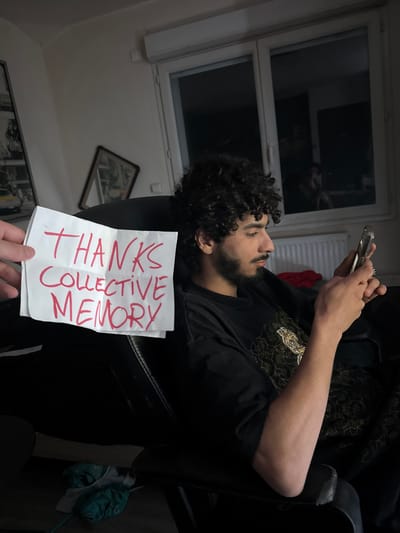 The image depicts an indoor scene in what appears to be a casual domestic setting, possibly in Marcoussis, France. A young man with dark, curly hair and a beard is lying on a dark armchair or sofa, horizontally across the frame. He is focused on a smartphone, which he holds with both hands. He is wearing a dark t-shirt with a decorative gold or yellow pattern across the chest and shoulders.

In the foreground, a hand holds up a white piece of paper. The paper has text written in bold red capital letters that reads: "THANKS COLLECTIVE MEMORY".

The background features a white-framed window, possibly an attic window, and a white radiator directly beneath it. The room's lighting is dim, suggesting it might be evening or night. On the wall, a framed picture is visible, tilted at an angle, and another framed item is partially seen towards the bottom of the frame. The overall atmosphere is relaxed and informal, with the man seemingly absorbed in his phone while a note is presented to him.