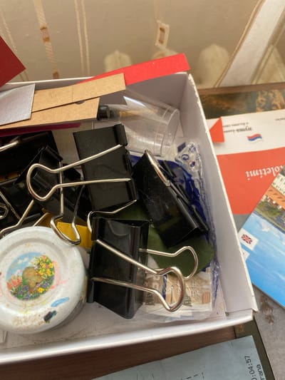 The image shows a close-up, high-angle view of an open white cardboard box filled with various craft supplies and office stationery. Dominating the frame are several large black binder clips with silver metal handles. Among these are also some pieces of colored paper, including red and tan, and a sheet of metallic silver paper. A small, round white object with a colorful illustration of a bee and flowers is partially visible on the left side, possibly a lid or a decorative element. Further in the box, a clear plastic cup and some plastic packaging are visible. In the background, a portion of a postcard or pamphlet featuring a city skyline and flags, including the Union Jack, is present. Another document with Cyrillic text and a stylized flag is also visible. The contents appear to be stored loosely within the box. The overall environment suggests a personal crafting or office space. The location of Bílina, Czechia is not depicted in the image.