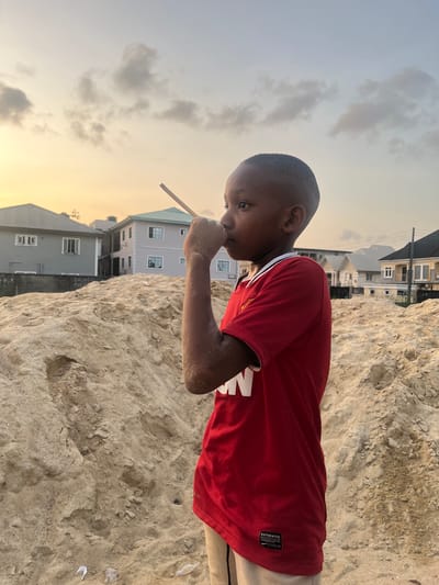 An outdoor scene captured at dusk or sunset in Iranla, Nigeria, depicts a young child of dark complexion, approximately 5-8 years old, standing in profile facing left. The child wears a red short-sleeved t-shirt with white trim and light-colored shorts. Their right arm and hand are visibly coated in light brown sand. The child's right hand is raised towards their mouth/nose area, holding a slender light-colored wooden stick between the thumb and index finger, extending upwards. The child's gaze is directed forward.
The foreground and midground are dominated by a large, irregularly shaped mound of light brown sand with visible indentations and disturbed areas. In the background, beyond the sand pile, several modern residential buildings are present under a sky transitioning from light blue to warm orange and yellow, with scattered grey clouds. Buildings include a grey two-story house with a dark roof on the left, and a purple/grey two-story house with a teal roof and external staircase in the center-right. Additional residential structures with varying roof colors are visible further right.