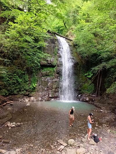 The image shows a waterfall in a lush, green forest. Two women are standing near the base of the waterfall, one in a black tank top and tan shorts, the other in a striped tank top and denim shorts. Both are wearing baseball caps. One woman is looking at her phone, while the other is looking at the waterfall. A black backpack and a red water bottle are on the ground near the woman in denim shorts. The waterfall is surrounded by large rocks and dense vegetation. The water is a clear, turquoise color, and the ground is covered in pebbles and rocks. The image appears to have been taken in the city of Plastunka, Russia, known for its natural beauty and waterfalls.