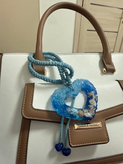 The image shows a close-up of a white leather tote bag with brown trim. A blue braided cord is looped through the handle, and a blue resin heart charm with small shells and white pebbles embedded in it hangs from the cord. Two small blue heart-shaped charms are attached to a blue string dangling below the heart charm. A rectangular gold-colored metal tag with the name "DANIELLE PATRICI" embossed on it is affixed to the front of the bag. The background is a plain, light-colored wall. The image does not provide any information about the location being Vladivostok, Russia, or any specific context beyond the objects depicted.