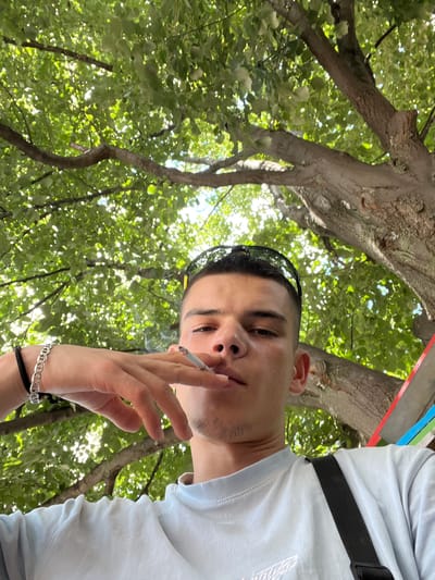 A young man with short dark hair and sunglasses perched on his head is taking a selfie outdoors. He is holding a cigarette in his right hand and appears to be exhaling smoke. He is wearing a light blue t-shirt and a black strap across his chest, possibly from a bag. The background is dominated by lush green foliage and tree branches, suggesting he is under a large tree. Sunlight filters through the leaves, creating dappled light. The man's left wrist is adorned with a silver chain bracelet and a black string bracelet. The image is shot from a low angle, looking up towards the man and the trees. The location is identified as Sopot, Bulgaria, implying an outdoor setting in that city.