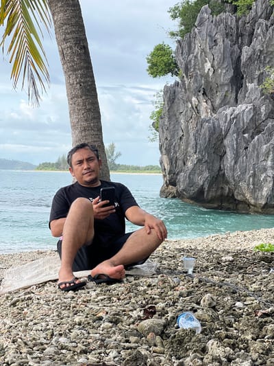A man sits by a palm tree on a beach in Ujongsedheun, Indonesia, holding a smartphone. He is wearing a black t-shirt and shorts. The beach is covered in pebbles and coral fragments. In the background, the turquoise water of the sea meets a rocky cliff face. The sky is overcast. There is a plastic bottle and a glass with a straw on the ground near the man.