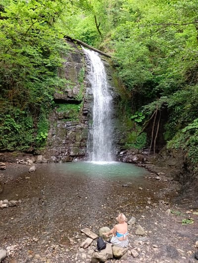The image shows a waterfall in a lush, green forest. A person is sitting on rocks by the water's edge, facing away from the camera. The waterfall cascades down a rocky cliff face, and the water collects in a clear, turquoise pool at the bottom. The surrounding area is dense with vegetation, including trees, bushes, and moss-covered rocks. The scene appears tranquil and natural.