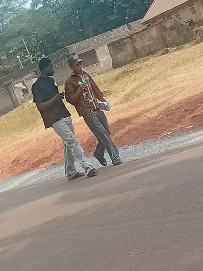 The image displays two males walking on a paved road in Ayangba, Nigeria. The male on the left, dressed in a black short-sleeved t-shirt and light grey baggy jeans, holds a small, light-colored rectangular object in his left hand and looks towards the male on the right. The male on the right wears a camouflage baseball cap, a brown long-sleeved shirt with a graphic design, and dark grey jeans. He holds a clear plastic bottle with a green cap in his right hand and a light blue rectangular object in his left hand, his mouth open, indicating speech. The foreground features a dark grey asphalt road, bordered by a strip of lighter gravel and a reddish-brown earth embankment with dry, yellowish-green grass. The background includes concrete block walls, some appearing unfinished, dense green foliage, and the corner of a building with a reddish-brown sloped roof.