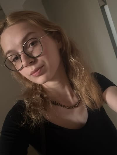 The image shows a close-up, overhead selfie of a young woman with blonde hair wearing glasses and a black top. She is wearing a beaded necklace. The background appears to be an indoor setting, possibly a room, with neutral-colored walls. There is no identifiable context within the image that would suggest a specific location in Kragujevac, Serbia.