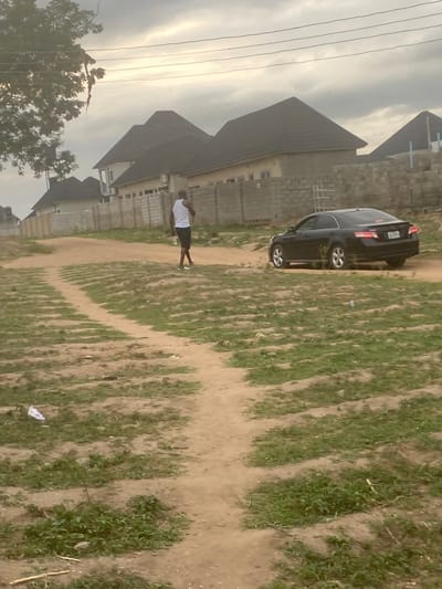 The image depicts an outdoor scene in Lugbe, Nigeria. A black car is parked next to a dirt path that is sparsely covered with grass. A person wearing a white shirt and dark shorts is standing on the path. Buildings with dark grey roofs are visible in the background, along with trees and utility lines. The sky is overcast.