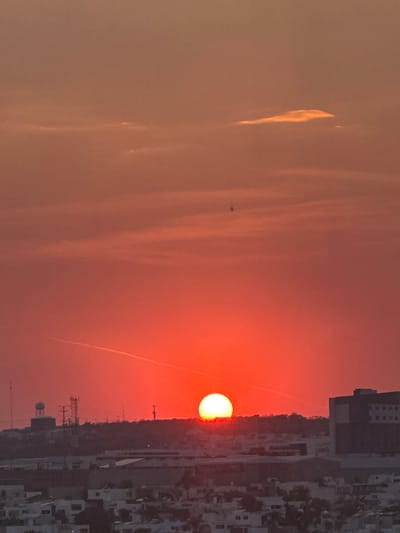 The image depicts a sunset in León, Mexico. The sun is a bright orange orb near the horizon, partially obscured by the silhouette of buildings. The sky transitions from a deep orange near the sun to a paler orange and grey higher up. There is a vapor trail from an airplane streaking across the sky, and a few wispy clouds are visible. In the foreground, there is an elevated building next to the rising sun. There are water towers and communication towers. The image has a slightly hazy or smoky quality, likely due to atmospheric conditions.