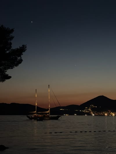 The image depicts a twilight scene at Sveti Stefan, Montenegro. In the foreground, a body of water is visible, with gentle ripples on its surface. A dark, silhouetted object, possibly a rock, is present in the lower left corner. A string of buoys stretches across the water in the mid-ground. A traditional wooden sailing vessel, illuminated by interior lights, is anchored on the water. Further in the distance, the silhouette of the island of Sveti Stefan and the mainland coastline is visible. The island and coastline are dotted with lights, suggesting a settlement. The sky transitions from a deep blue at the top to a warm orange and yellow hue near the horizon. Two celestial bodies, likely planets or bright stars, are visible in the upper portion of the sky. A dark, silhouetted tree branch extends from the upper left corner of the frame. The overall atmosphere is serene and evocative of a coastal evening.