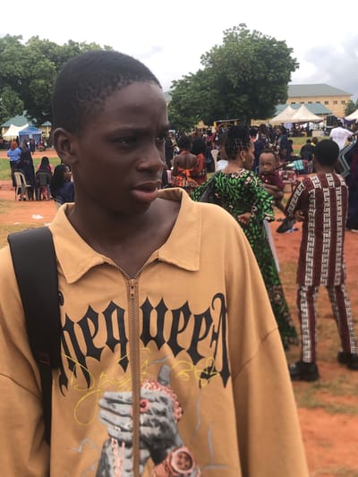 The image shows a close-up of a young dark-skinned person wearing a tan jacket with black Gothic-style writing and a print of hands with jewelry. The person is facing slightly to the left with a serious expression. In the background, there is a large crowd of people, trees, and a building or tent. The location is in Nsugbe, Nigeria. People are dressed in various outfits including a green and patterned dress, and a red and white patterned suit. The environment appears to be an outdoor gathering or event.