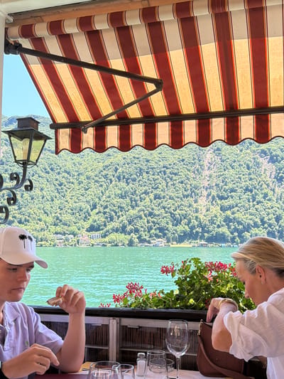 The image depicts a young person wearing a white baseball cap with palm trees and a light purple polo shirt, seated at a table. The person is holding a piece of food in their right hand, bringing it towards their mouth. To the right, an adult woman with blonde hair is also seated at the table, reaching into a brown purse. Both individuals are positioned in the foreground, with a table set with glasses, a salt shaker, and a napkin between them.
Behind them, a vibrant green lake stretches out, with a densely forested hillside rising on the opposite shore, dotted with buildings. Above the lake and hillside, a clear blue sky is visible. The scene is partially covered by a striped awning, with red and white bands, supported by dark metal poles. A decorative, black metal lamp with a light bulb is attached to the left side of the awning structure. The overall setting suggests an outdoor dining area overlooking a scenic lake and mountainous landscape, likely in Lugano, Switzerland, given the visual cues of the lake, surrounding vegetation, and architecture. The lighting indicates it is daytime.