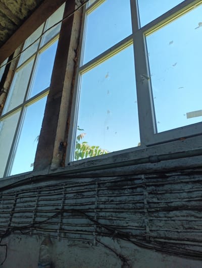The image depicts an interior view looking out a multi-paned window. The window frame appears to be white and is segmented into multiple panes. Some panes have small, irregular pieces of tape or debris attached to the glass. Through the windows, a clear blue sky is visible, with the upper branches and leaves of a tree partially obscuring the view in some sections.

The interior side of the window shows structural elements that appear weathered and worn. A thick, brown, beam-like structure runs vertically along the left side of the window frame. The wall below the window is textured and shows a pattern of horizontal lines, possibly indicating a building material or a conduit system. Several dark electrical wires are routed across this textured wall, with some appearing to be secured by clips. A partially visible plastic bottle with a blue cap rests on the floor in the lower left corner.

The scene suggests an industrial or abandoned building, possibly undergoing renovation or in a state of disrepair. The lighting indicates it is daytime, with sunlight streaming through the window. The overall impression is one of an unkempt, utilitarian space. No people are visible in the image. The location is identified as the city of Karnobat, Bulgaria.