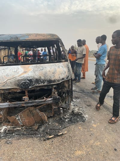 The image captures the aftermath of a vehicle fire in Alizaga, Nigeria, showing a heavily burnt-out bus or large van on a paved road. The vehicle, charred black and grey, appears skeletal, with its body panels and interior extensively damaged by flames. Twisted metal, exposed wiring, and ash are visible within its structure and on the road surrounding it.
A group of approximately ten men and boys, mostly young adults, are gathered around the vehicle, observing the damage. They are dressed in casual attire, including t-shirts, jeans, and traditional clothing. Some individuals are standing closer to the burnt wreck, looking inside or at its remains, while others stand further back. Additional, indistinct figures can be seen inside the burnt vehicle's frame.
The scene is set outdoors during daylight hours, under a hazy or overcast sky, which contributes to a somewhat muted color palette. The road surface is asphalt, and the background shows an open, relatively flat landscape. No discernible text is visible in the image. The expressions of the onlookers are not clearly visible, but their presence indicates curiosity or concern following the incident. The burnt debris on the road surface emphasizes the intensity of the fire.
