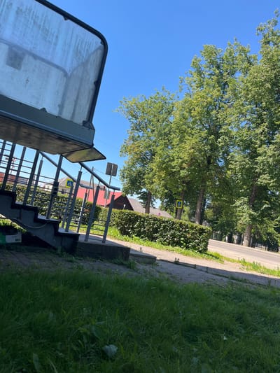 The image shows a bright, sunny day in what appears to be a suburban or semi-rural setting in Ludza, Latvia. The scene is dominated by lush green trees and grass, suggesting a temperate climate and a time of year with abundant vegetation, likely summer.

In the foreground, a portion of a structure with a transparent or frosted covering is visible on the left. To the right, a metal staircase with railings leads downwards, hinting at an elevated walkway, bridge, or perhaps an entrance to a building. The steps themselves are made of dark material, possibly concrete.

Beyond the staircase, a well-maintained green hedge runs parallel to a paved pathway. Further in the background, several mature trees line a road. A distinctive yellow and blue pedestrian crossing sign is mounted on one of the trees, indicating a designated area for people to cross the road. Red-roofed buildings are partially visible behind the hedge and trees, suggesting residential or public structures. The road itself is paved and appears to be relatively quiet.

The overall impression is one of a peaceful, well-kept outdoor environment on a clear, sunny day. There is no visible text other than the symbol on the pedestrian crossing sign. No people are present in the image, and the scene appears to be static, with no specific activity taking place. The strong sunlight casts distinct shadows, further confirming the time of day is likely late morning or early afternoon.