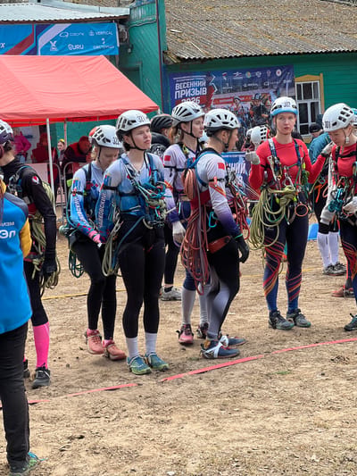 The image shows a group of climbers standing in a line, apparently preparing for a competition in Lesnoye, Russia. They are wearing climbing helmets and harnesses with ropes attached. Each is wearing tight-fitting athletic clothing, some with racing numbers attached to their helmets, in a dirt field marked by a pink tape line. There is a banner with the words "ВЕСЕННИЙ ПРИЗЫВ" ("Spring Call") and logos of sponsors such as "OZONE", "VERTICAL", and "GURU" visible. A partially visible orange tent and building structures are visible in the background.