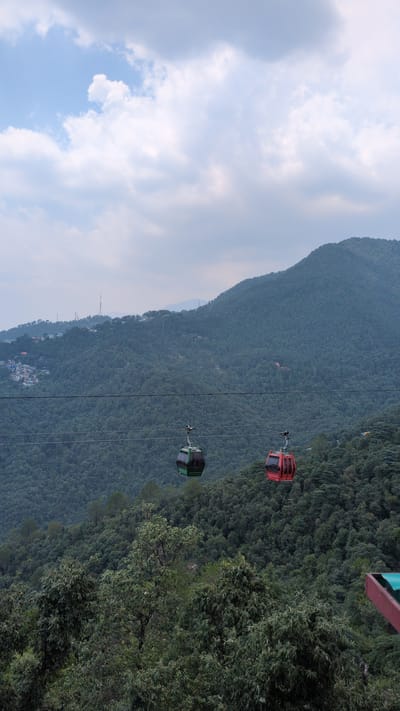 This is a photograph of a cable car system in Dharamshala, India. The cable cars are traveling over a lush, densely forested mountainside. Two cable cars are visible in the image, one green and one red. The green cable car is to the left and the red cable car is to the right. In the background, the mountain is covered with trees and some buildings are visible on the slope. The sky is cloudy with patches of blue. The cable cars appear to be moving. The image is taken from a perspective below the cable cars, looking up towards the mountain and the sky.