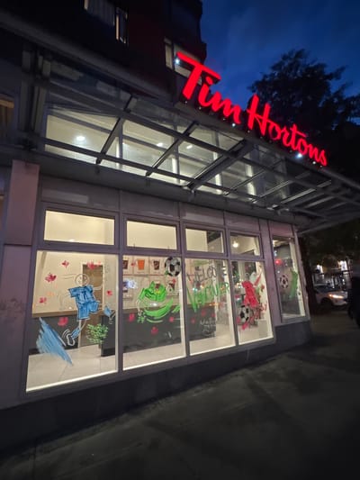 The image shows a Tim Hortons storefront at dusk. The iconic red neon Tim Hortons sign is prominently displayed above the entrance. The storefront windows are decorated with colorful drawings, including figures of people, soccer balls, and maple leaves, suggesting a festive or celebratory theme. There are also images of beverages on display inside. The windows are illuminated from within, casting a warm glow onto the street outside. The street in front of the store is paved, and a sidewalk is visible. In the background, a white car and a few blurred figures of people can be seen, indicating a public urban setting. The overall atmosphere is lively and welcoming, despite the dim lighting of the evening. The location is identified as Vancouver, Canada, based on the text on the window which says "WELCOME VANCOUVER".