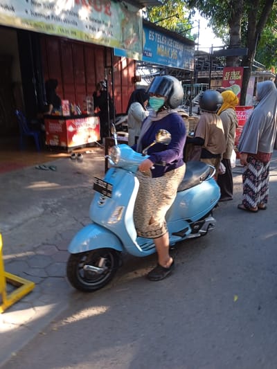 The image shows a woman wearing a helmet and a face mask on a light blue scooter, likely in Madiun, Indonesia. Several other women stand nearby. Some wear head coverings, and one wears a patterned skirt. Behind them are storefronts and food stands, including one selling "Es Teler Ice Cream." Other people are in the background. The scooter is parked on a paved surface. The light suggests it is daytime.