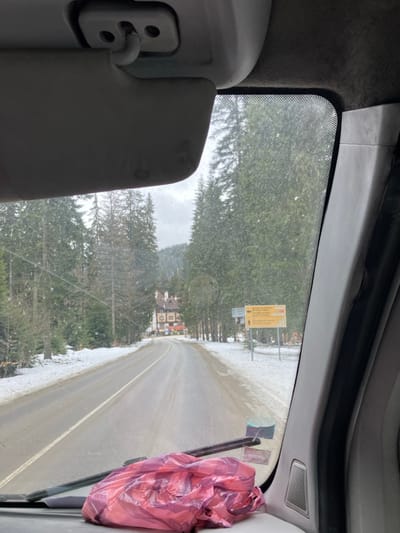 The image provides a forensic view from inside a vehicle, looking forward through the windshield onto a winter road scene in Stoikite, Bulgaria. In the immediate foreground, a grey sun visor is visible in the upper left, and a crumpled pink plastic bag rests on the dashboard, partially obscuring the windshield wiper and lower portion of the windshield.

The scene outside features a two-lane asphalt road, partially cleared of snow, which curves gently to the left and ascends into the mid-distance. Snow covers the verges of the road and the surrounding terrain. On both sides of the road, dense evergreen forests, primarily composed of tall pine trees, line the landscape, extending up the slopes of visible hills or mountains under an overcast sky.

In the mid-distance, positioned to the left of the road, a multi-story building with a brown roof and light-colored facades (possibly a hotel or lodge) is partially visible through the trees. To the right of the road, a yellow directional sign with multiple arrows and text in what appears to be Bulgarian and English is visible in the snow-covered roadside, indicating directions to various locations or facilities. There are no individuals, explicit actions, or interactions depicted within the frame.
