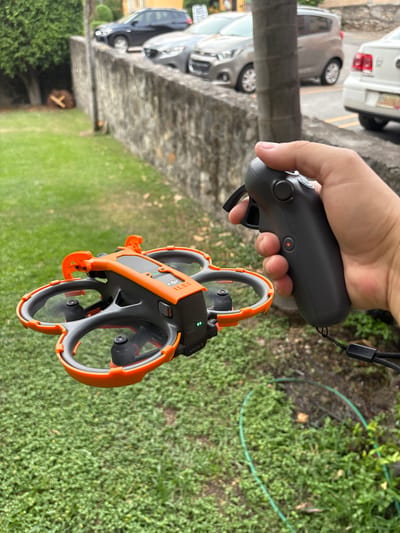 A hand holds a black remote control, connected to a black and orange drone. The drone is above a patch of green grass. In the background, a row of parked cars is visible along a stone structure, and trees are visible in the distance. The image was taken in Cuernavaca, Mexico.