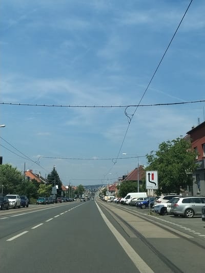 The image depicts a street scene in Pilsen, Czechia, on a clear, sunny day. The perspective is from the driver's seat of a vehicle moving forward on a multi-lane road. The road has tram tracks running along the right side, with a line of parked cars to the right of the tracks. On the left side of the road, there are more parked cars and buildings lining the street. The buildings are a mix of residential and commercial properties, with some featuring red tile roofs. Trees and streetlights are also visible. A traffic sign showing a lane merge ahead, with a distance of 1700 meters, is positioned on the right side of the road. The sky is a bright blue with some wispy clouds. The overall impression is a typical urban street in a European city.