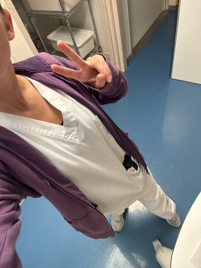 A person, presumed to be the photographer, is depicted in a high-angle, self-portrait photograph. They are wearing white pants and a white collared shirt, possibly scrubs, and a purple jacket. The person is making a peace sign with their left hand.
The background consists of a blue tiled floor and white shelves holding white rectangular containers. The environment appears to be an indoor space, possibly within a medical or industrial setting.
While the location of Tourcoing, France is mentioned in the instructions, there is no contextual information present in the photograph to verify or confirm this location.