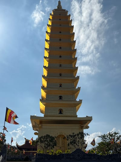 A tall, multi-tiered pagoda dominates the scene, viewed from a low angle and appearing almost vertical. The pagoda is beige and gold in color, with distinct horizontal sections and repeating architectural details. The sky is visible behind the pagoda, exhibiting a gradient from light blue to deeper blue with scattered clouds. Buddhist flags are visible near the base of the pagoda, adding to the architectural setting. The location is Diên Khánh, Vietnam, as determined by the pagoda's architectural style, the presence of Buddhist flags, and the region's typical environmental characteristics.