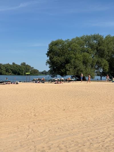 The image shows a sandy beach with people sunbathing and walking. In the background, there is a lake or river with trees and a clear blue sky. There are several lounge chairs on the sand, and some people are lying on them. One person is walking towards the water, and another person is standing near the water. There is a sign in the background that says "ПРОКАТА" which means "rental" in Russian. The scene appears to be a recreational area, possibly a beach resort. The location is identified as Zhdanovichsky Selsoviet, Belarus.